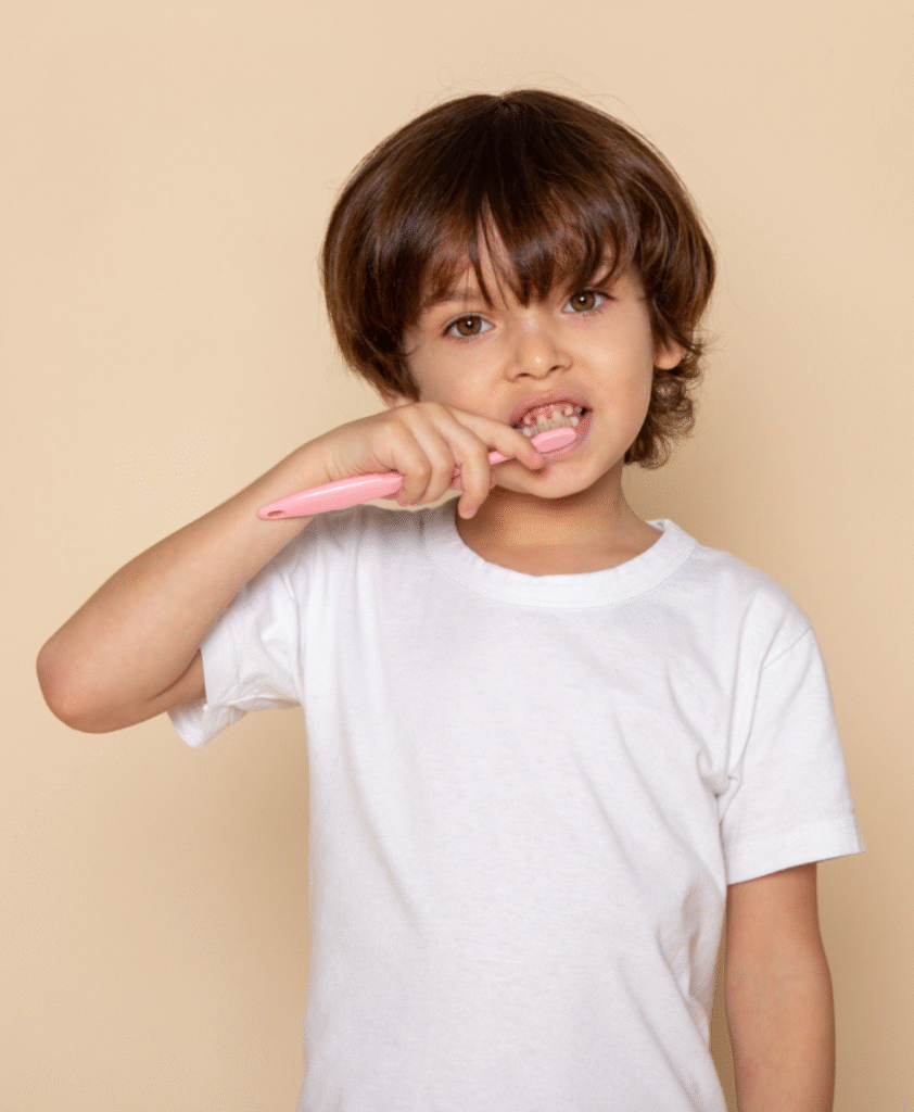 Малко момче с бяла тениска, което се усмихва, докато си мие зъбите с розова четка – дентална хигиена при деца