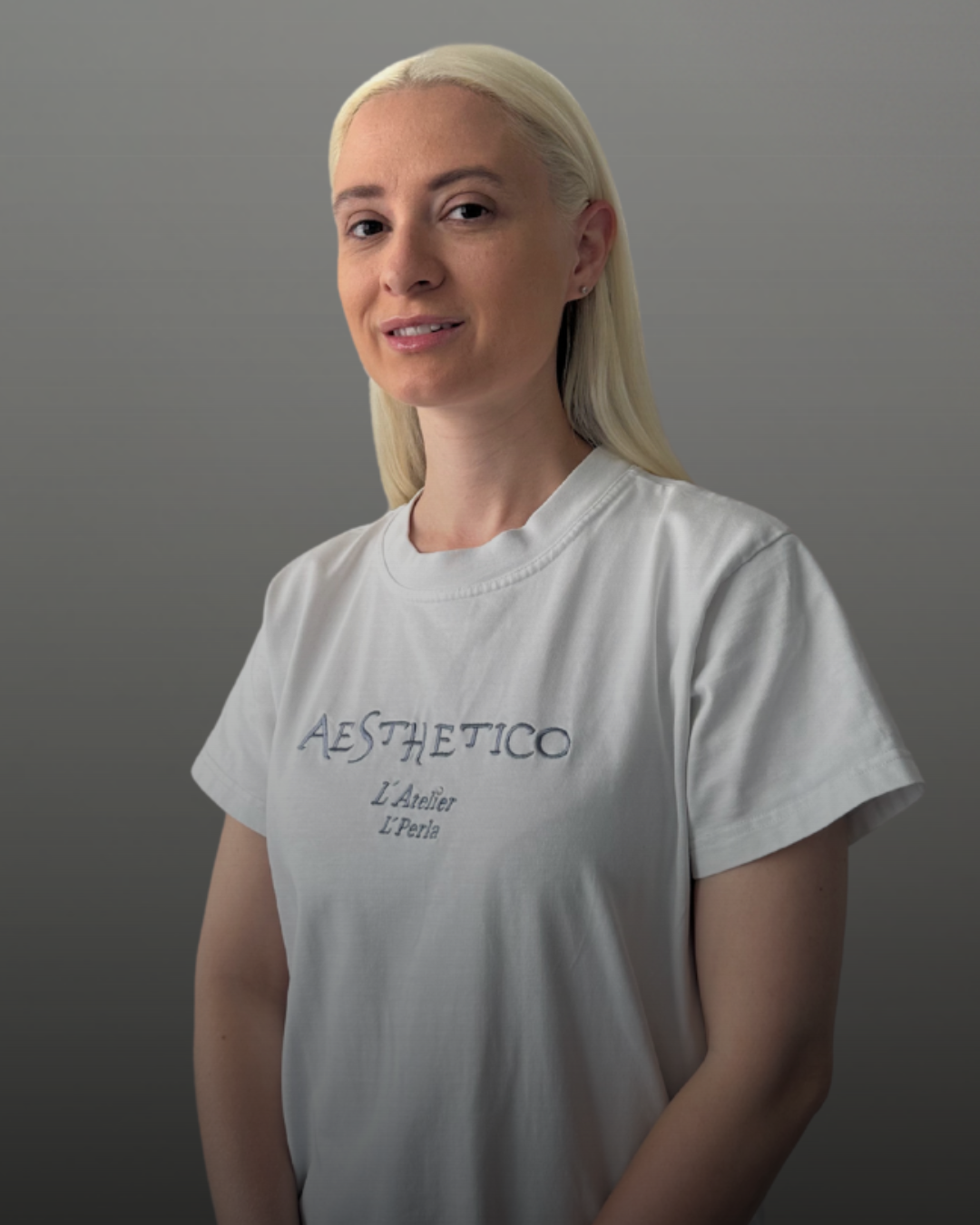 Портрет на лекар по дентална медицина с руса коса и бяла тениска с лого Aesthetico, на сив фон.