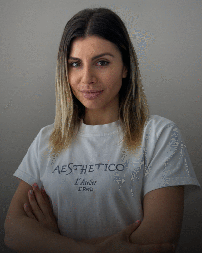 Портрет на дентална асистентка със светла коса и бяла тениска с лого Aesthetico, заснета на сив фон.