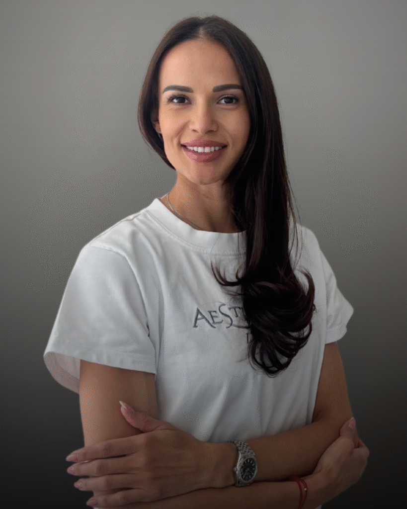 Портрет на дентална асистентка с дълга тъмна коса, облечена с бяла тениска с лого Aesthetico, на неутрален фон.