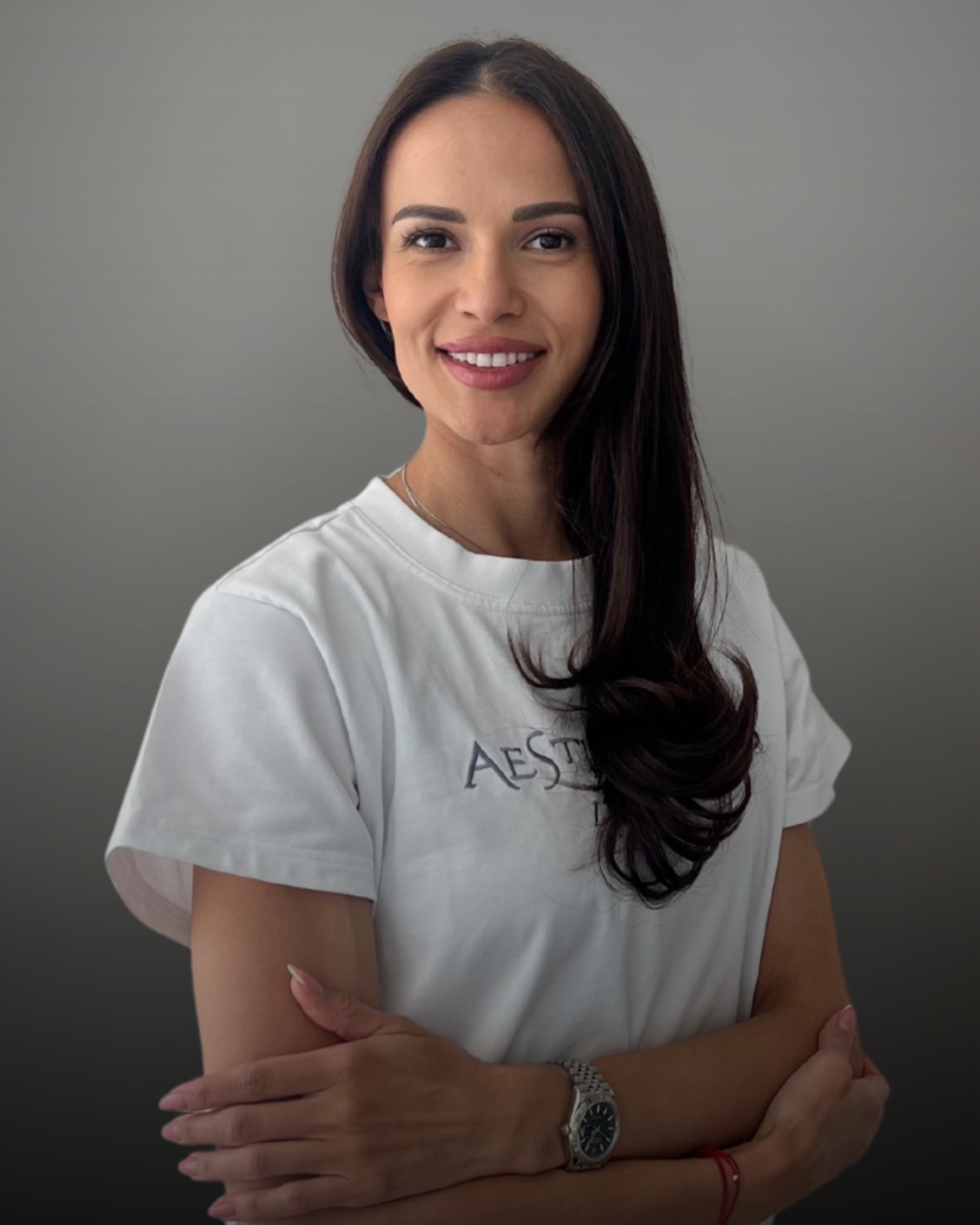 Портрет на дентална асистентка с дълга тъмна коса, облечена с бяла тениска с лого Aesthetico, на неутрален фон.